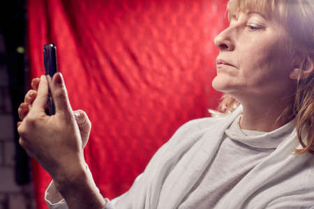 Portrait of a middle-aged woman with cell phone on a red background. Unprofessional female model doing selfie in the room or in the Studioの写真素材