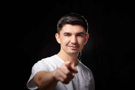 A young man who is a medical doctor in a white uniform poses against a black background in the...
