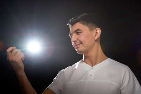 A young man who is a medical doctor in a white uniform poses against a black background in the...