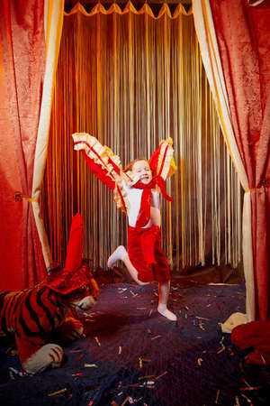 Small girl during a stylized theatrical circus photo shoot in a beautiful red location. Young model posing on stage with curtainの写真素材