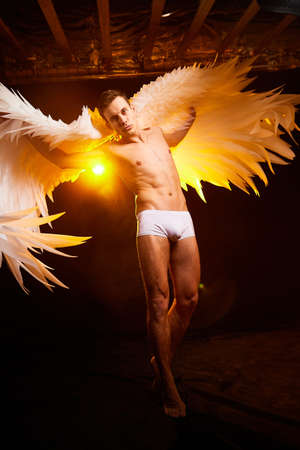 Handsome young athletic man with a bare torso who looks like an angel with white wings. Model dancer posing in a dark studio on black backgroundの写真素材