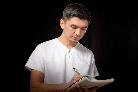 A young man who is a medical doctor in a white uniform poses against a black background in the studio with a notepad in handの写真素材