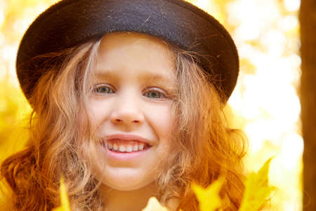 Portrait of young funny girl with blonde curly hair and in black hat in an autumn park on a yellow and orange leaf backgroundの写真素材