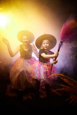 Beautiful brunette mother and cute little daughter looking as witches in special dresses and hats in room decorated for Halloween. Witchcraft and wizardry in carnival. Halloween style photo shoot.の写真素材