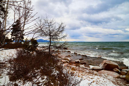 Nature landscape with dramatic sky and clouds, lake with ice, shore with snow and dark trees in cold winter or autumn dayの写真素材
