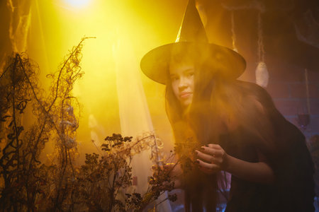 Young girl looking like witch having fun on Halloween in a dark room with yellow light and smoke....