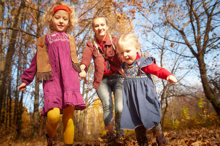 Two young little girl and his mother with blonde hair in an autumn park on a yellow and orange leaf background. Family walking in forestの写真素材