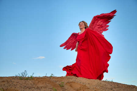 A plump blonde middle-aged woman with red angle wings in a desert with dunes and sand in a nice summer or spring sunny day with blue skyの写真素材