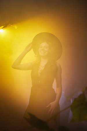 Young slim woman looking like witch having fun on Halloween in a dark room with yellow light and...