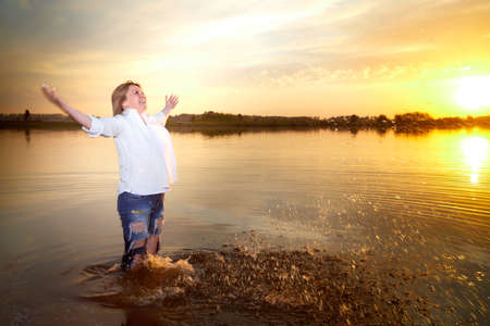 Portrait of middle-aged woman near lake or river during beautiful sunset with sun above the horizon and yellow reflection in the waterの写真素材