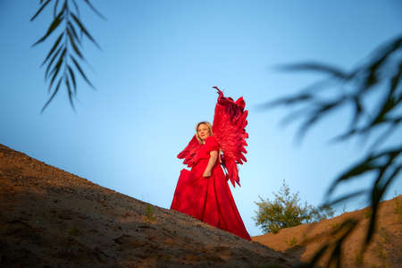 A plump blonde middle-aged woman with red angle wings in a desert with dunes and sand in a nice summer or spring sunny day with blue skyの写真素材