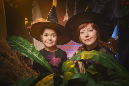 Mom and daughter in costumes for a holiday of halloween in dark decoration and with nice yellow light. Funny nice family during carnival in the roomの写真素材