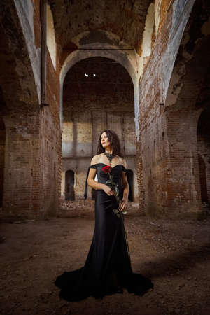 A brunette girl with long hair in an evening dress in a vintage Gothic castle. A woman who looks like an evil witch or sorceress in an abandoned old church. Female Model posing in Halloweenの写真素材