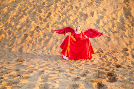 A plump blonde middle-aged woman with red angle wings in a desert with dunes and sand in a nice...