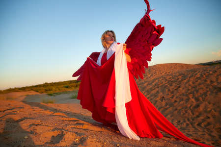 A plump blonde middle-aged woman with red angle wings in a desert with dunes and sand in a nice summer or spring sunny day with blue skyの写真素材