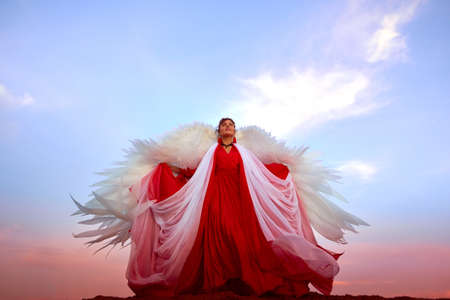 Beautiful young woman or girl with curly hair and in red dress with a light flying fabric and white wings on the sand on sunny day with blue sky. Angel model or dancer posing in photo shoot on dunesの写真素材