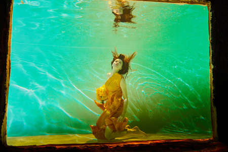 Girl in a beautiful yellow dress under water behind of glass. Female model posing under water in swimming poolの写真素材