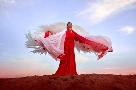 Beautiful young woman or girl with curly hair and in red dress with a light flying fabric and white wings on the sand on sunny day with blue sky. Angel model or dancer posing in photo shoot on dunesの写真素材