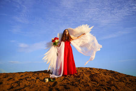 Beautiful young woman or girl in red dress and white wings on the sand on sunny day with blue sky. Angel model or dancer posing in photo shoot on dunesの写真素材