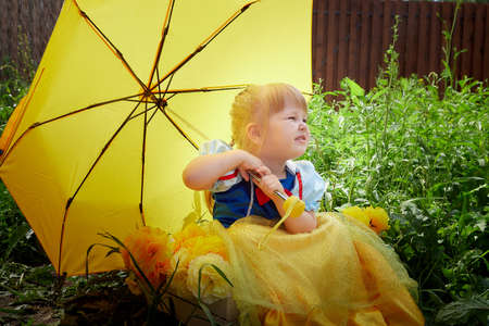 A beautiful pretty little girl looking like a doll in a beautiful dress with yellow umbrella in nature on a summer dayの写真素材