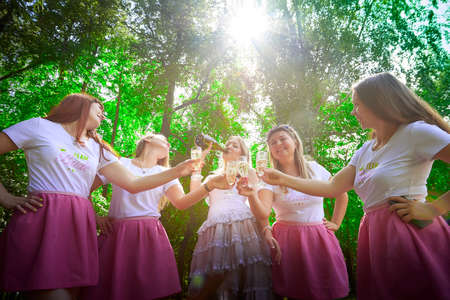 Kirov, Russia - July 29, 2021: Bride with bridesmaids in the same short pink skirts with a bottle of alcohol and glasses in green park in summerのeditorial素材
