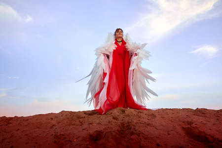 Beautiful young woman or girl with curly hair and in red dress with a light flying fabric and white wings on the sand on sunny day with blue sky. Angel model or dancer posing in photo shoot on dunesの写真素材