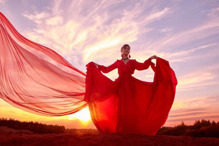 Beautiful young woman or girl with curly hair and in red dress with a light flying fabric on the sand on sunny day with blue sky on background. Model or dancer posing in photo shoot on sand dunesの写真素材