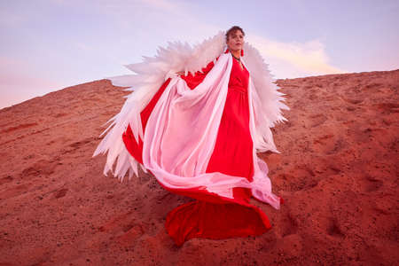 Beautiful young woman or girl with curly hair and in red dress with a light flying fabric and white wings on sand on a sunny day with blue sky. Model or dancer posing in photo shoot on dunesの写真素材