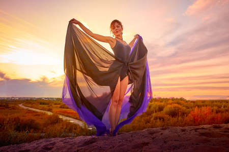Beautiful girl with black dress and violet fluttering cloth dancing on the sand dune. Model or dancer posing in nature landscapeの写真素材