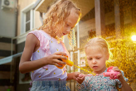 Two cute little blonde girls sisters with soap bubbles under rain drops near house and village in a summer sunny day. Family having rest and fun in good spring timeの写真素材