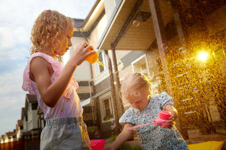 Two cute little blonde girls sisters with soap bubbles under rain drops near house and village in a summer sunny day. Family having rest and fun in good spring timeの写真素材