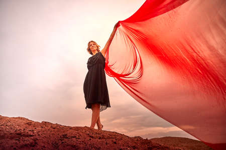A girl in black dress with red fabric dances on sand dunes against a dramatic sky before a thunderstorm. Model posing during photo shoot on natureの写真素材