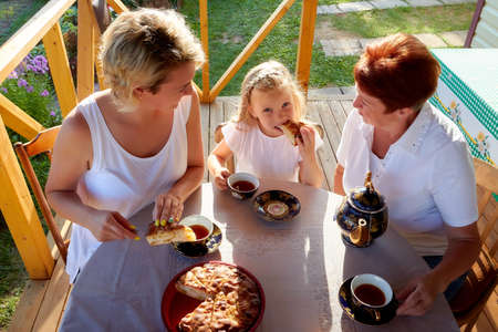 Little blonde girl, mother and grandmother at a table on wooden terrace on nature or in the village...