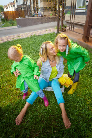 Two cute little blonde girls sisters in green raincoats and mother on a green lawn under rain drops in a summer sunny day near rural house in a village. Family having rest and fun in good spring dayの写真素材
