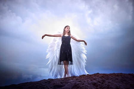 Young slim girl in black dress with with white wings dances on sand dunes against a dramatic sky before a thunderstorm. Model posing during photo shoot on nature. Beautiful angel in rain dayの写真素材