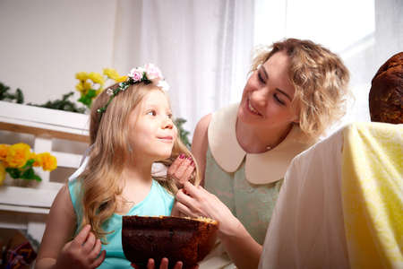 Nice mom and beautiful blonde daughter in room durin spring photo shoot before easter with festive bread, kulesh or cake. Happy girl and woman having joy and eating togetherの写真素材