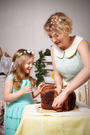 Nice mom and beautiful blonde daughter in room durin spring photo shoot before easter with festive bread, kulesh or cake. Happy girl and woman having joy and eating togetherの写真素材