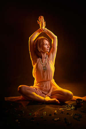 A beautiful adult skinny blonde woman in a beautiful light dress does yoga exercises on a black...