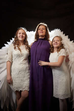 Mother with curly hair and cute teenagers girls with white wings looking like a nice angels posing in photo shoot in studio with black background. Family having fun and joy togetherの写真素材