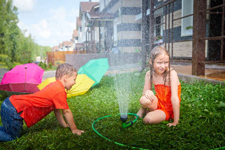 Children playing with garden sprinkler. Brother and sister running and jumping. Summer outdoor water fun in backyard. Boy and girl play with hose watering grass. Kids run and splash on hot sunny dayの写真素材