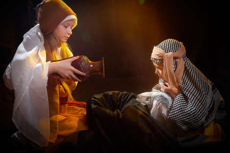 Little skinny girl and small boy in long white and yellow dress having fun with water and jug. Young model sister and brother in dark studio in stylized Arabic east costume of Israel or Palestineの写真素材
