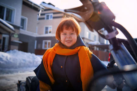 Stylish happy kid girl 2-3 year old with redhair wear blue coat and yellow scarf walk in city street in winter, autumn or spring season with snowの写真素材