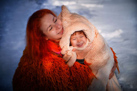 Mother with red hair and fluffy orange jacket and frozen little daughter in a warm blanket walking in winter, autumn or spring in forest. The concept of love and friendship of mom and girlの写真素材