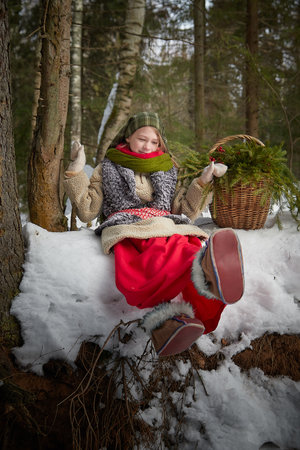 Nice cute teenager girl in old peasant clothes with hot fur coat and shawl in cold winter forest with fir trees, pines and snow on winter day. Child with basket full of green branches and red berriesの写真素材