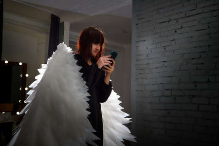 Young elegant woman in trendy black clothes with white wings. Brunette model in nice dress posing in studio in fashion lookbook as angel with cell phone. Alone girl in dark room takes a selfieの写真素材