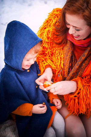 An extraordinary mother with red hair and a bright fluffy jacket and a cute little daughter in a fashionable stylish coat with an orange in the forest. Family woman and girl walking in winterの写真素材