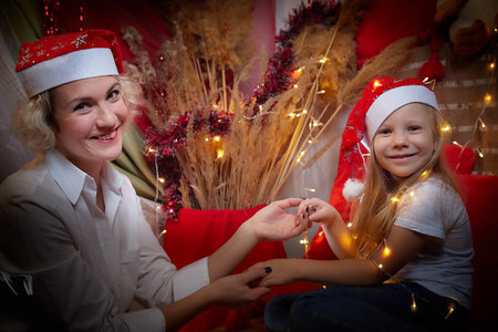 Cute mother and daughter in the red hats of Santa Claus assistants in a room decorated for Christmas. The tradition of decorating house and dressing up for holidays. Happy childhood and motherhoodの写真素材