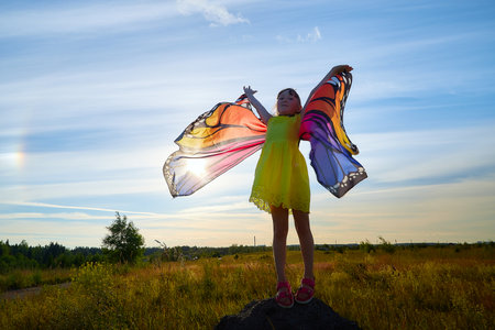 Beautiful little girl in yellow dress and in colorful bright butterfly wings in field with green grass, yellow flowers and blue sky in a sunny summer or spring dayの写真素材