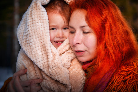 Mother with red hair and fluffy orange jacket and frozen little daughter in a warm blanket walking in winter, autumn or spring in forest. The concept of love and friendship of mom and girlの写真素材