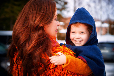 Mother with red hair and a fluffy orange jacket and little daughter with yellow flowers walking in winter, autumn or spring in village and snow. The concept of love and friendship of mom and girlの写真素材